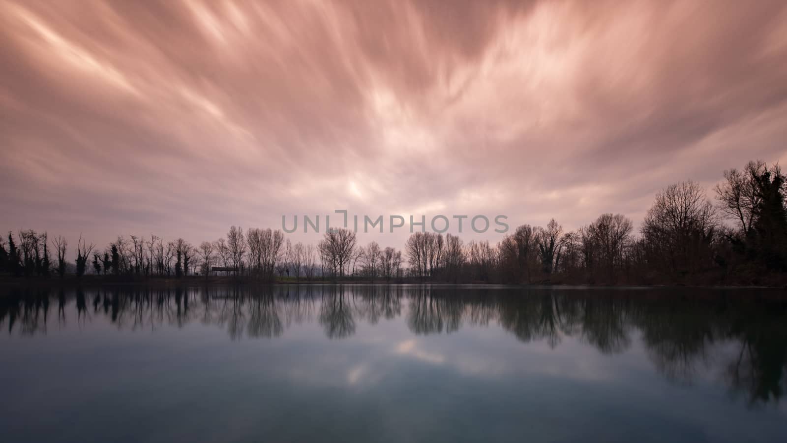 A red and cloudy sky reflected in the water of a lake at sunset  by brambillasimone
