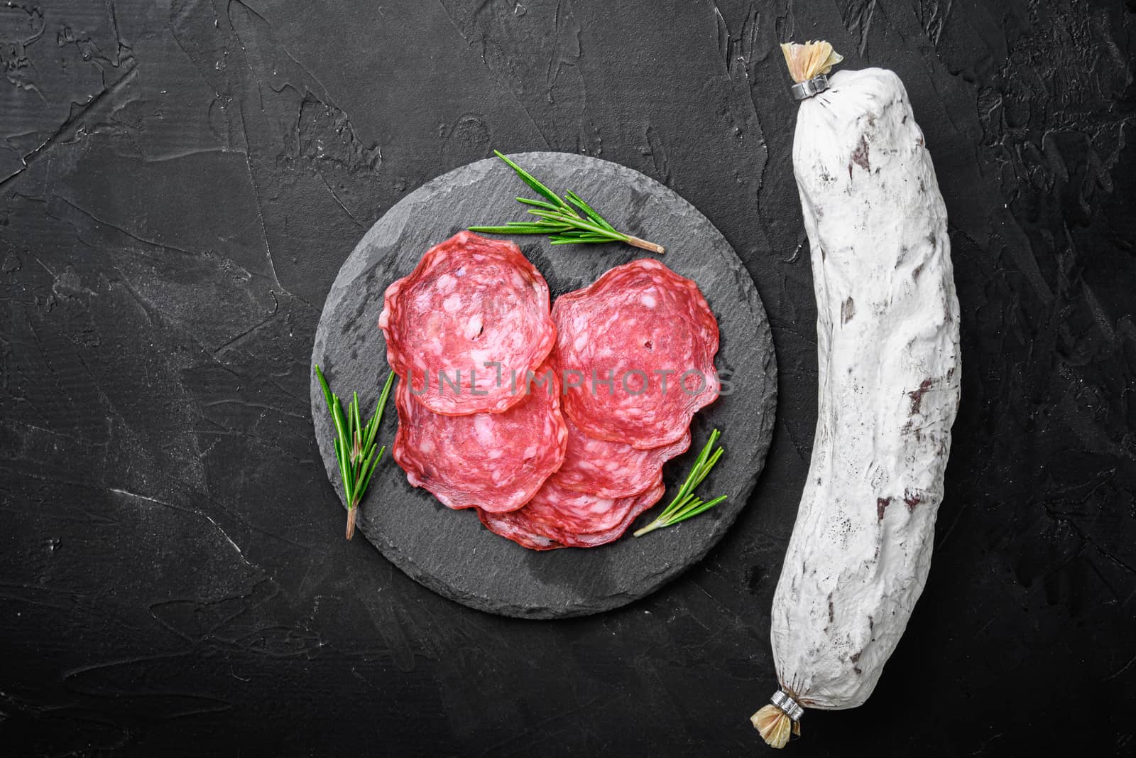 Dry cured, whole and slices cuts of salchichon sausage on black textured surface, topview with space for text.