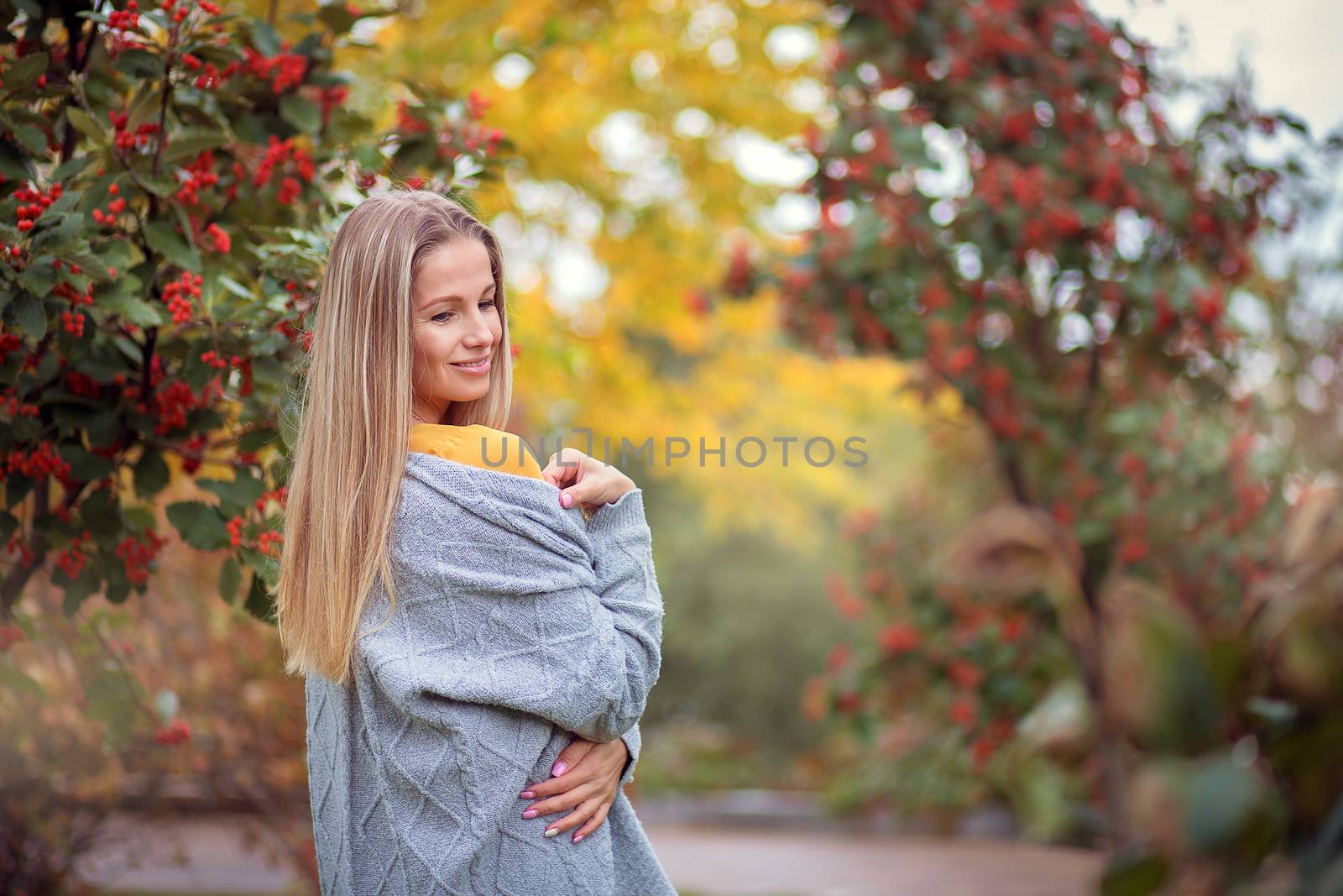 Blonde girl in a gray cardigan and a yellow dress among the autumn trees with red berries. Autumn theme