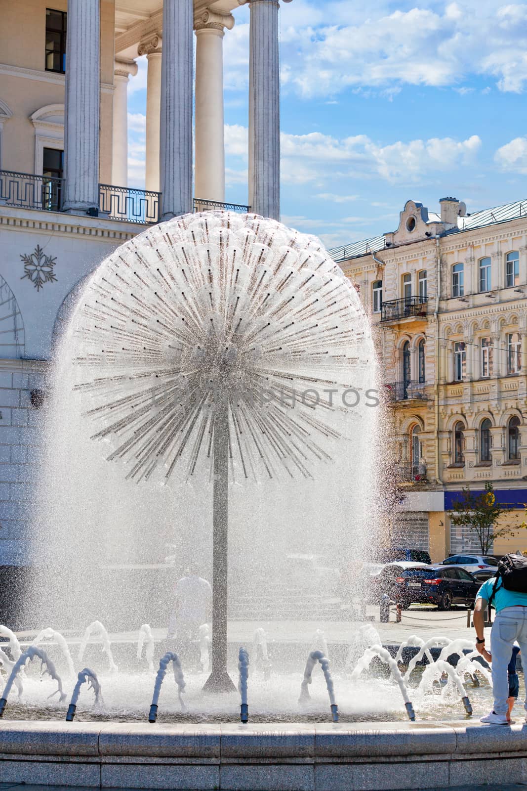 A beautiful city fountain in the form of a huge dandelion refreshes people against the background of the arched-column facade of the Kyiv City Conservatory on a sunny summer day. by Sergii