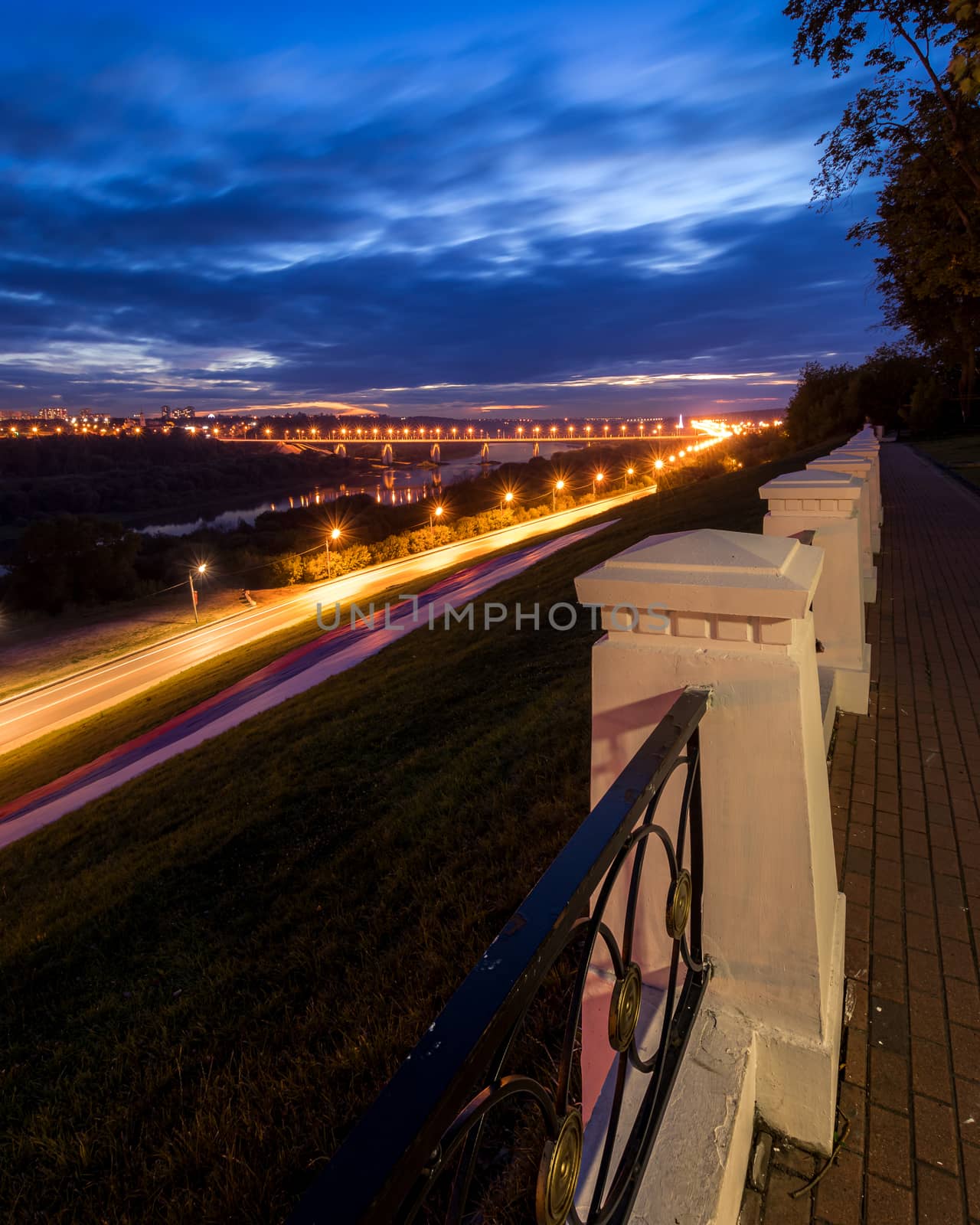 Moving car with blur light through city at night. Bridge over the river and the road. A view from the park from a height with a fence in the foreground. Cityscape.