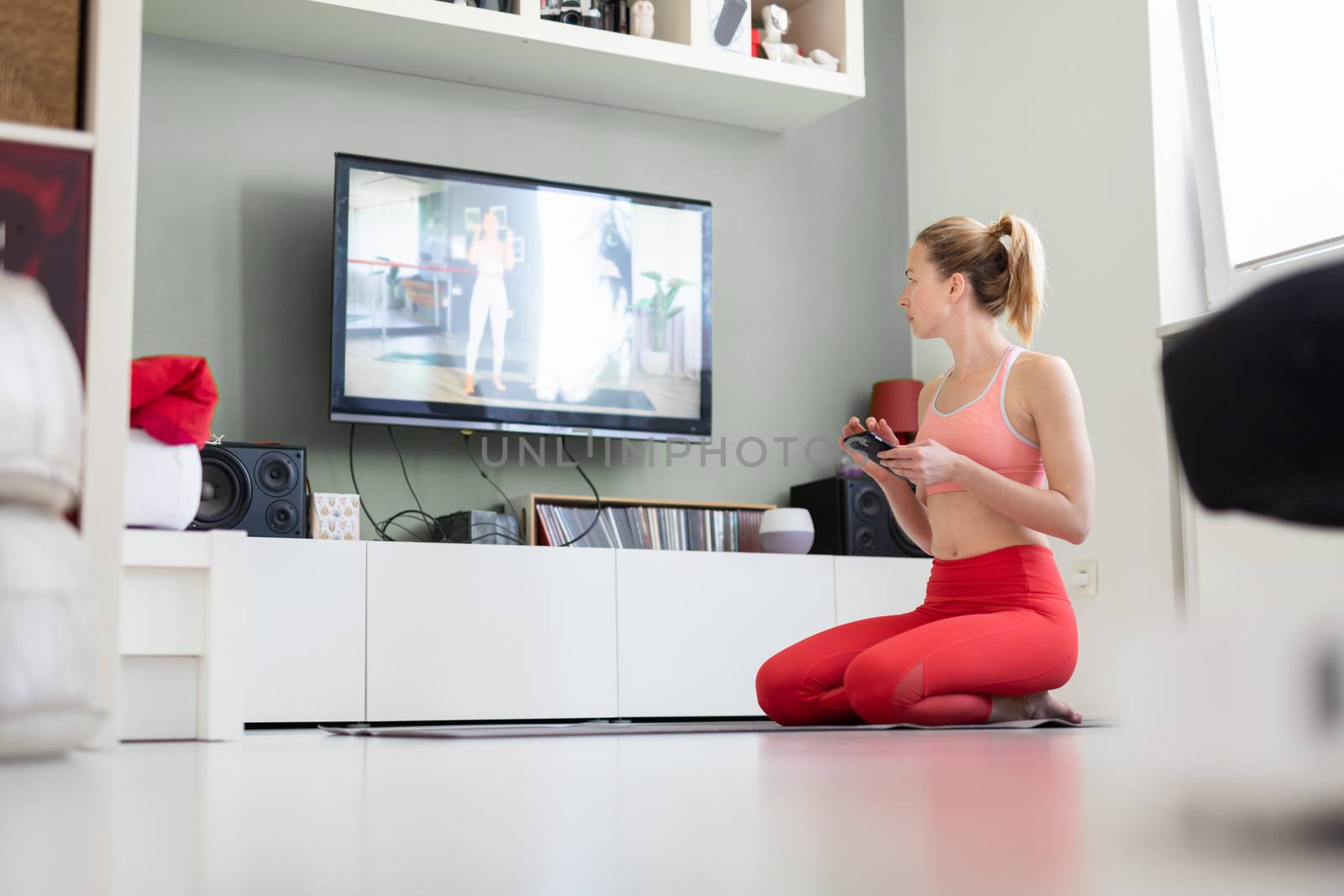 Attractive sporty woman working out at home, doing pilates exercise in front of television in her small studio appartment. Social distancing. Stay healthy and stay at home during corona virus pandemic by kasto