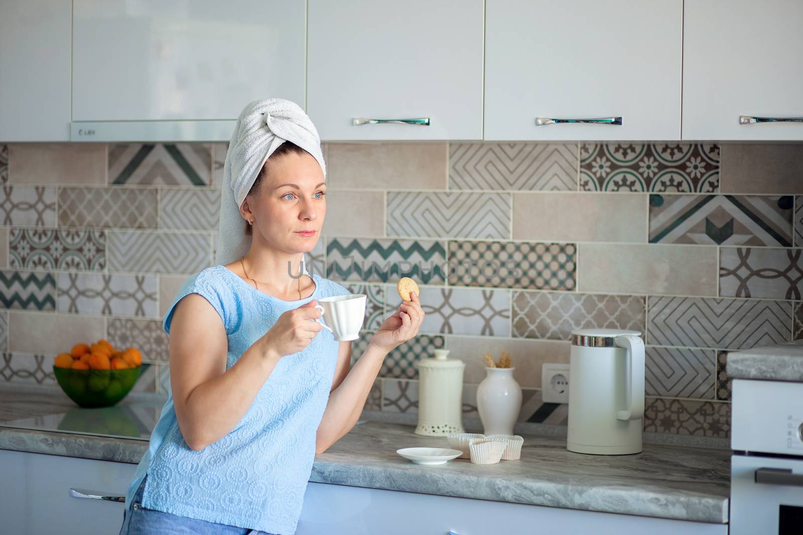 Happy woman with a towel on her head sutra drinks coffee with cookies for breakfast in his own kitchen at home by borisenkoket