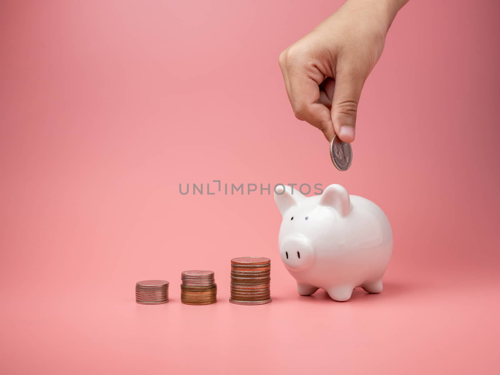 White piggy Bank and coin and human hand putting coin in piggy bank on a pink background. Copy space for design.