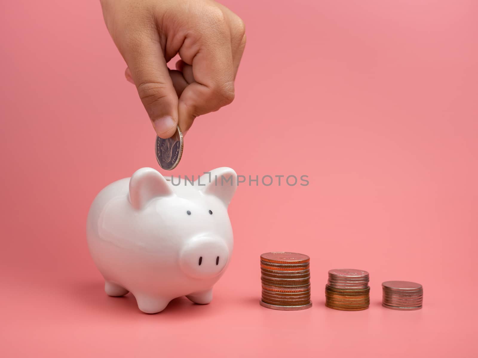 White piggy Bank and coin and human hand putting coin in piggy bank on a pink background. Copy space for design.