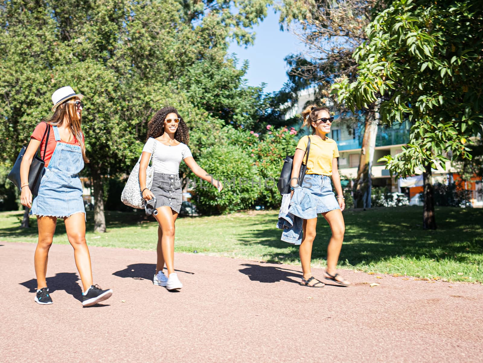 Three young mixed race female friends walking together in the park smiling with sunglasses - Two Caucasian women take a fun walk in the nature in the city with a Hispanic woman in casual wear by robbyfontanesi