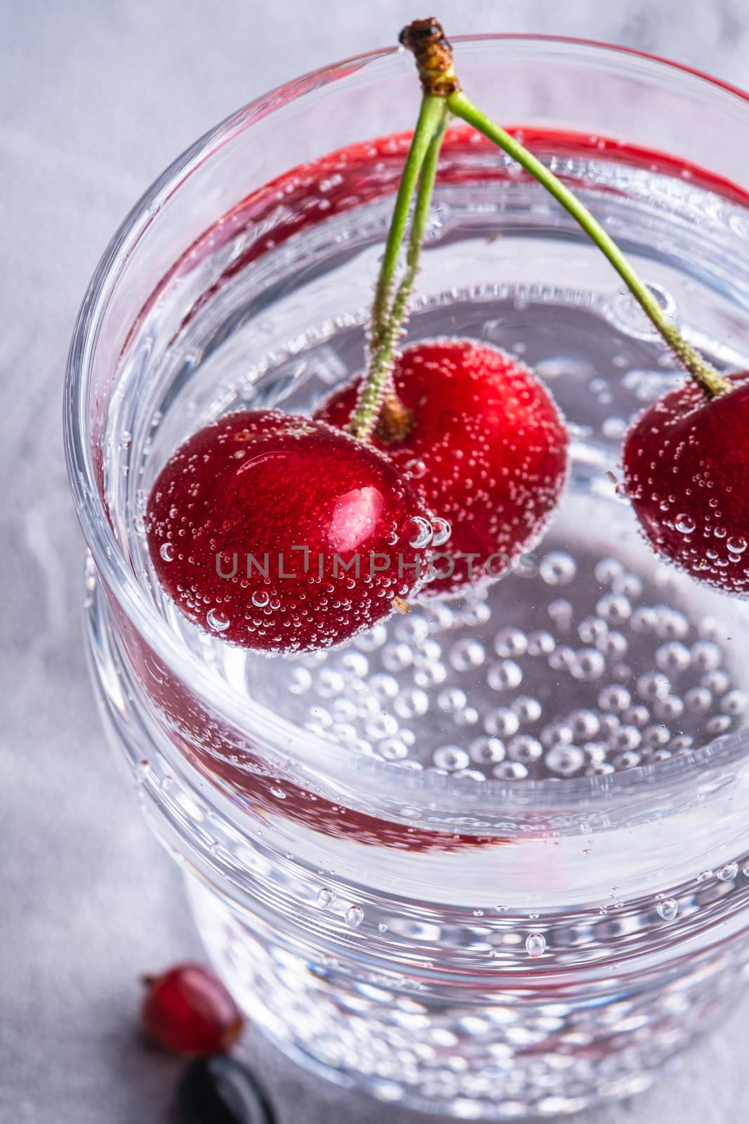 Fresh cold sparkling water drink with cherry, raspberry and currant berries in transparent glass on stone concrete background, summer diet beverage, angle view macro