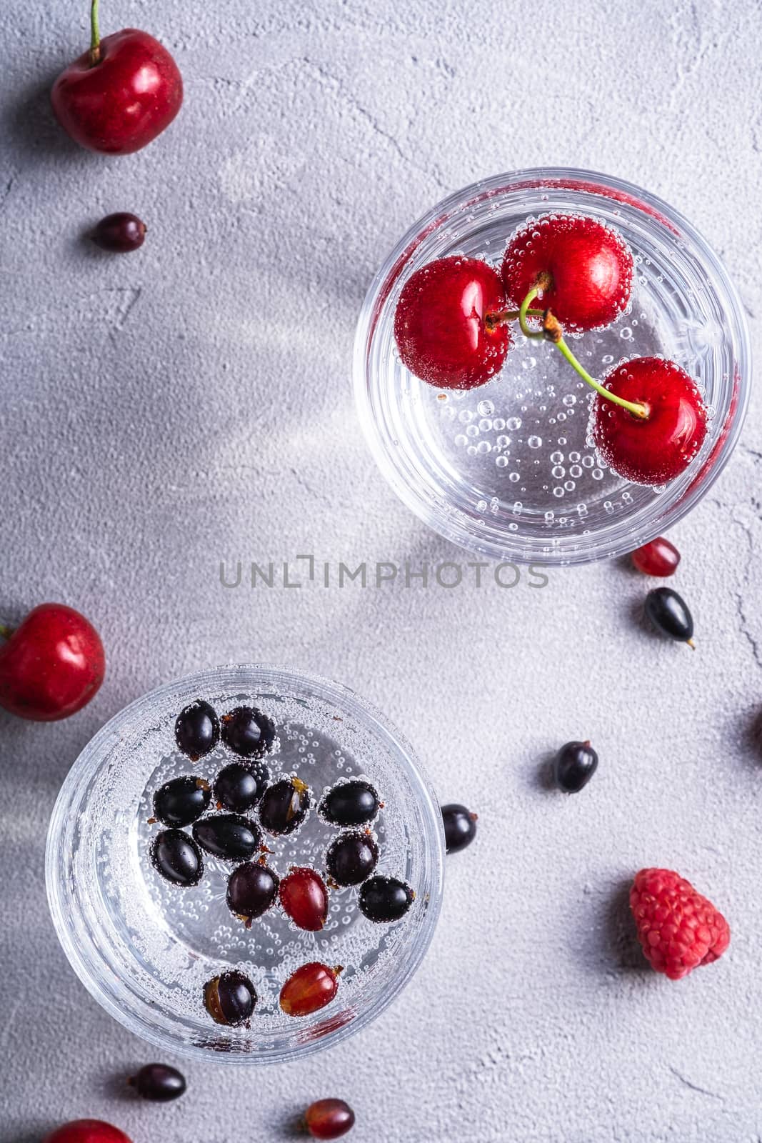 Fresh cold sparkling water drink with berries in two transparent glass on stone concrete background, summer diet beverage, top view