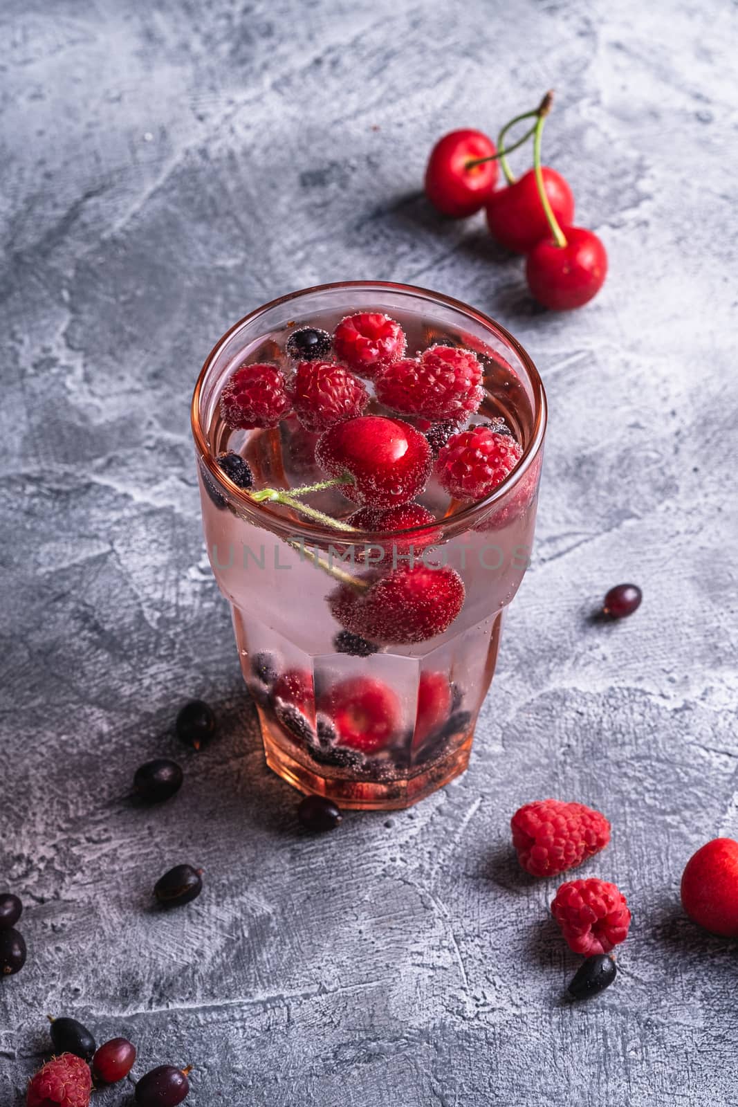 Fresh cold sparkling water drink with cherry, raspberry and currant berries in red faceted glass on stone concrete background, summer diet beverage, angle view