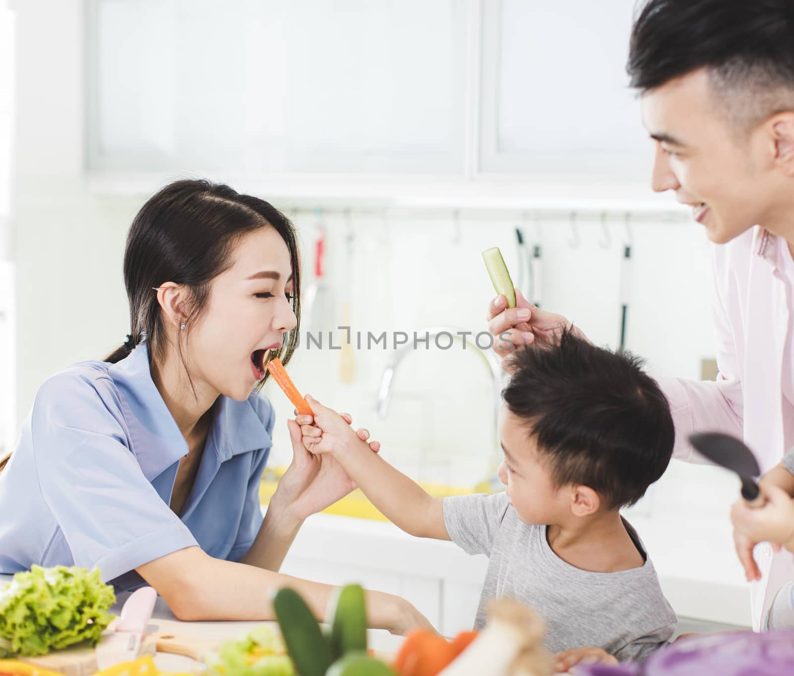 happy little boy feeding mother a piece of carrot in kitchen by tomwang