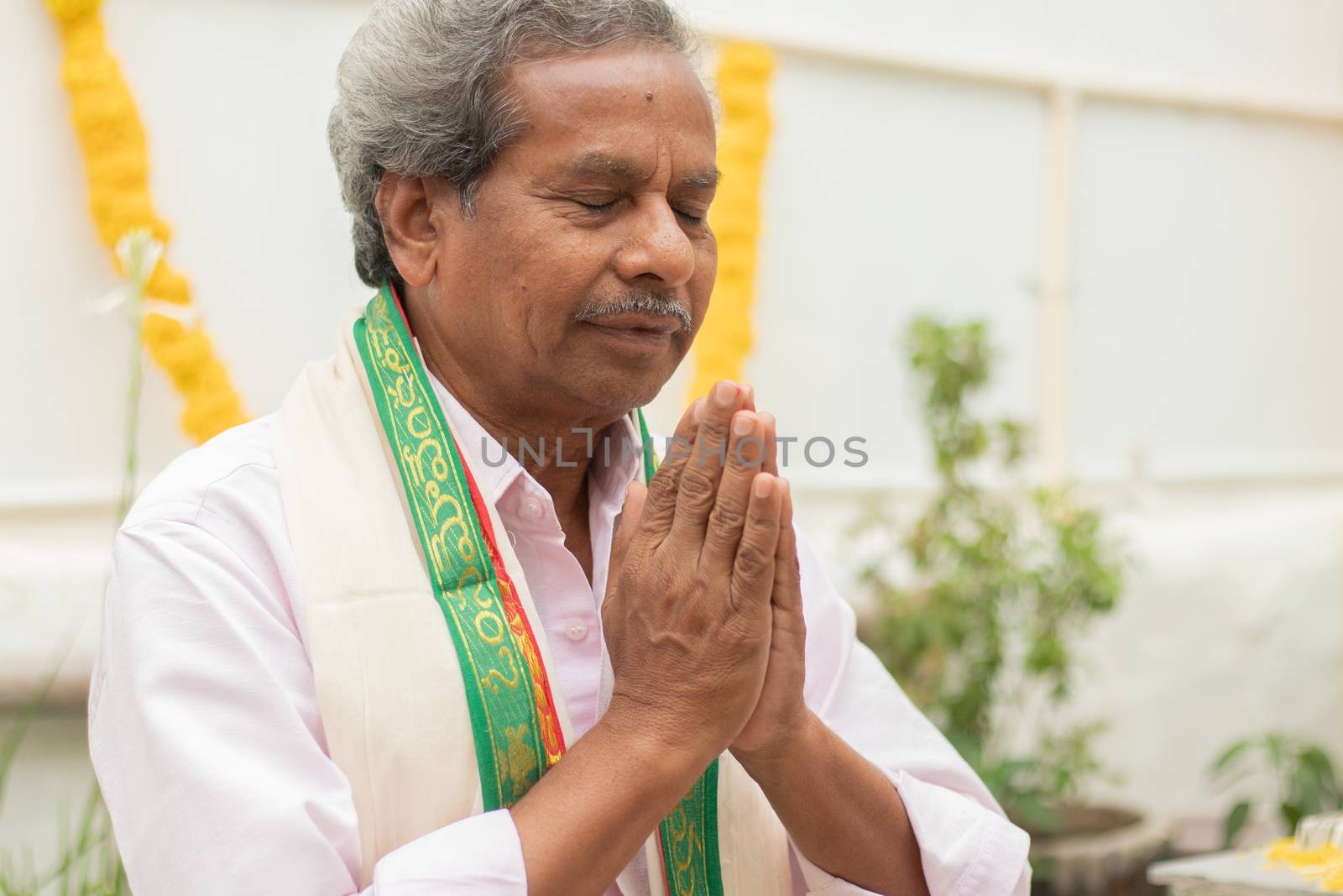 Elder man in closed eyes offering Prayer by namaste gesture praying to god at home in traditional dress during rituals