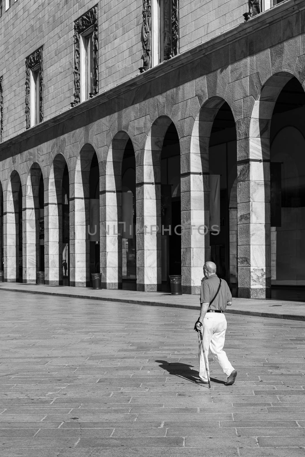 An elderly man with walking stick walks through the streets of the city of Milan, black and white street photography