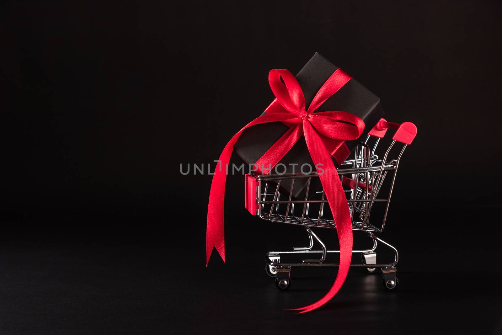 Black Friday sale shopping concept, Gift box wrapped black paper and red bow ribbon present in the shopping cart, studio shot on black background