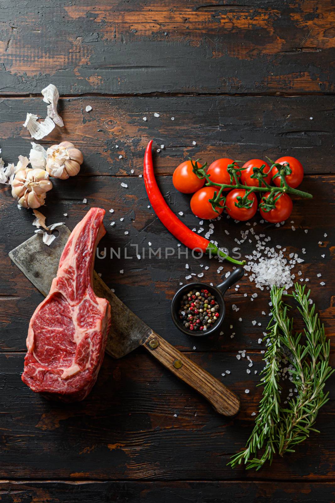 Raw dry steak cowboy on dark wooden vintage table background. Tomahawk steak or Scotch Fillet BBQ with seasonings, rosemary, tomato, chikie, garlic, peppercorn, salt with meat cleaver knife Top view. Free space for text. by Ilianesolenyi
