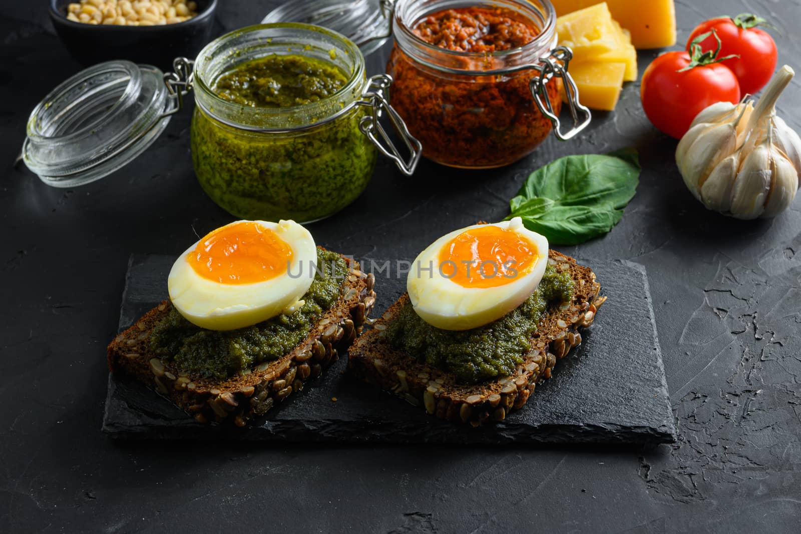 red and green Pesto alla Genovese , Basil Sauce resh home made and cooking italian recipe ingredients Parmesan cheese, basil leaves, pine nuts, olive oil, garlic, salt, tomatoes chulkboard side view on black stone concrete surface close up.
