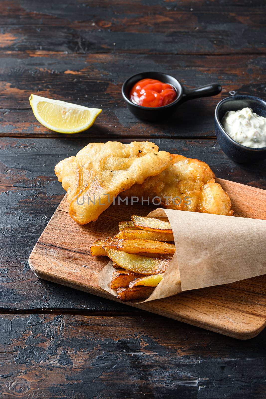 British Traditional Fish and chips with mashed peas, tartar sauce inn crumpled paper cone on wood chopping board dip and lemon - fried cod, french fries, lemon slices, tartar sauce, ketchup tomatoe served in the Pub or Restaurant over old wooden planks dark table side view close up by Ilianesolenyi