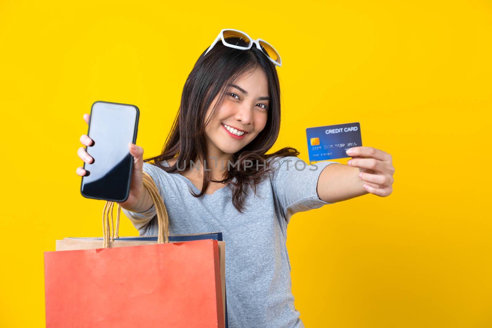 Happy Asian smiling woman presenting credit card and mobile phone with Carrying a shopping coloful bag for presenting online shopping on isolated yellow color background, copy space and studio