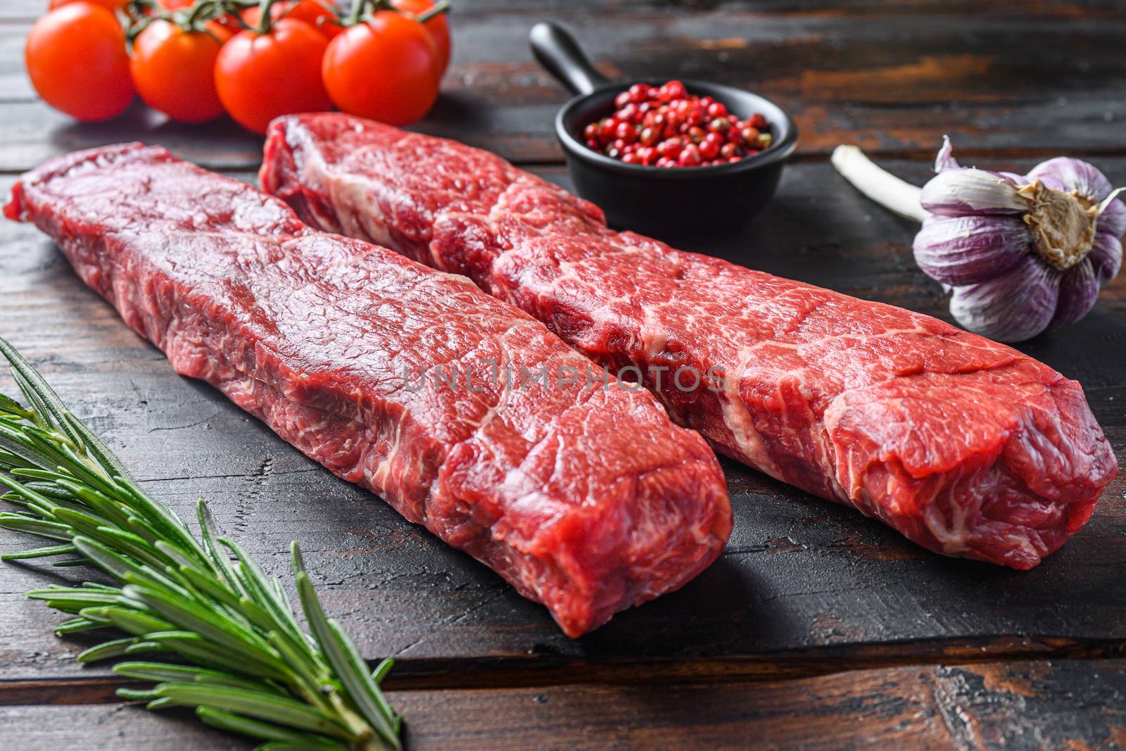 Two fresh, raw steaks from the cut of beef called the denver steak or organic top blade wuth herbs on old dark wooden table surface side view close up.