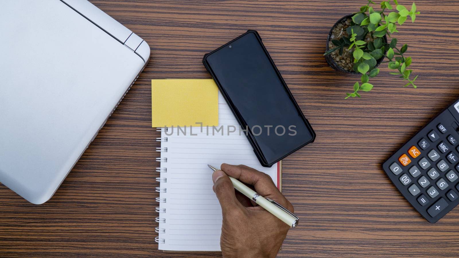 Writing on a notepad while working from home. A lap top, a mobile, calculator and plant are also on display on this brown striped working table. by sonandonures