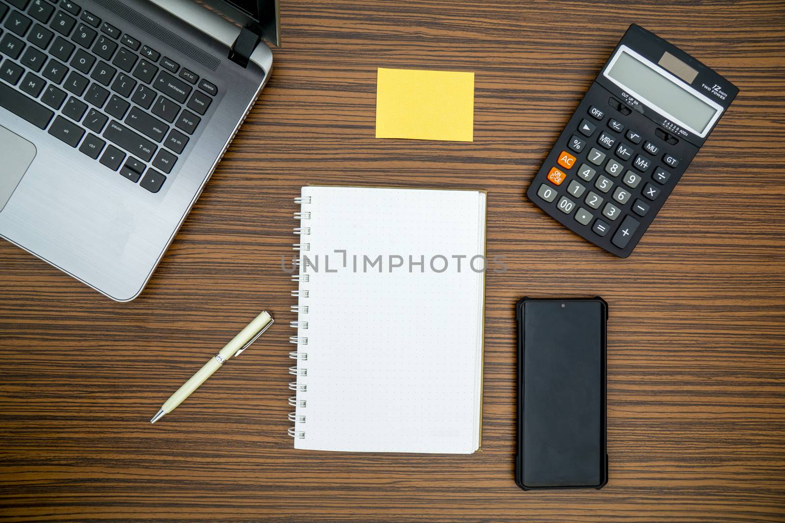 An office table working enviroment. Notepad, sticky note, pen plant, calculator and a lap top on a brown striped zebrawood design table top. Must have objects while working from home during Covid-19 by sonandonures