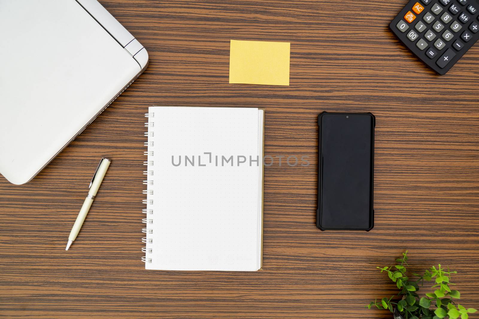 An office table working enviroment. Notepad, sticky note, pen plant, calculator and a lap top on a brown striped zebrawood design table top. Must have objects while working from home during Covid-19