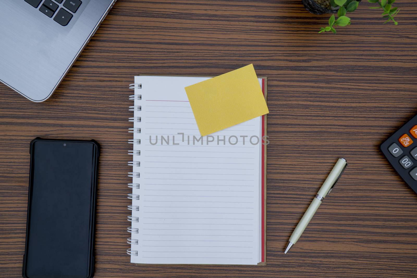 Striped brown working table with note pad, mobile phone, pen, calculator a laptop and table plant on display.