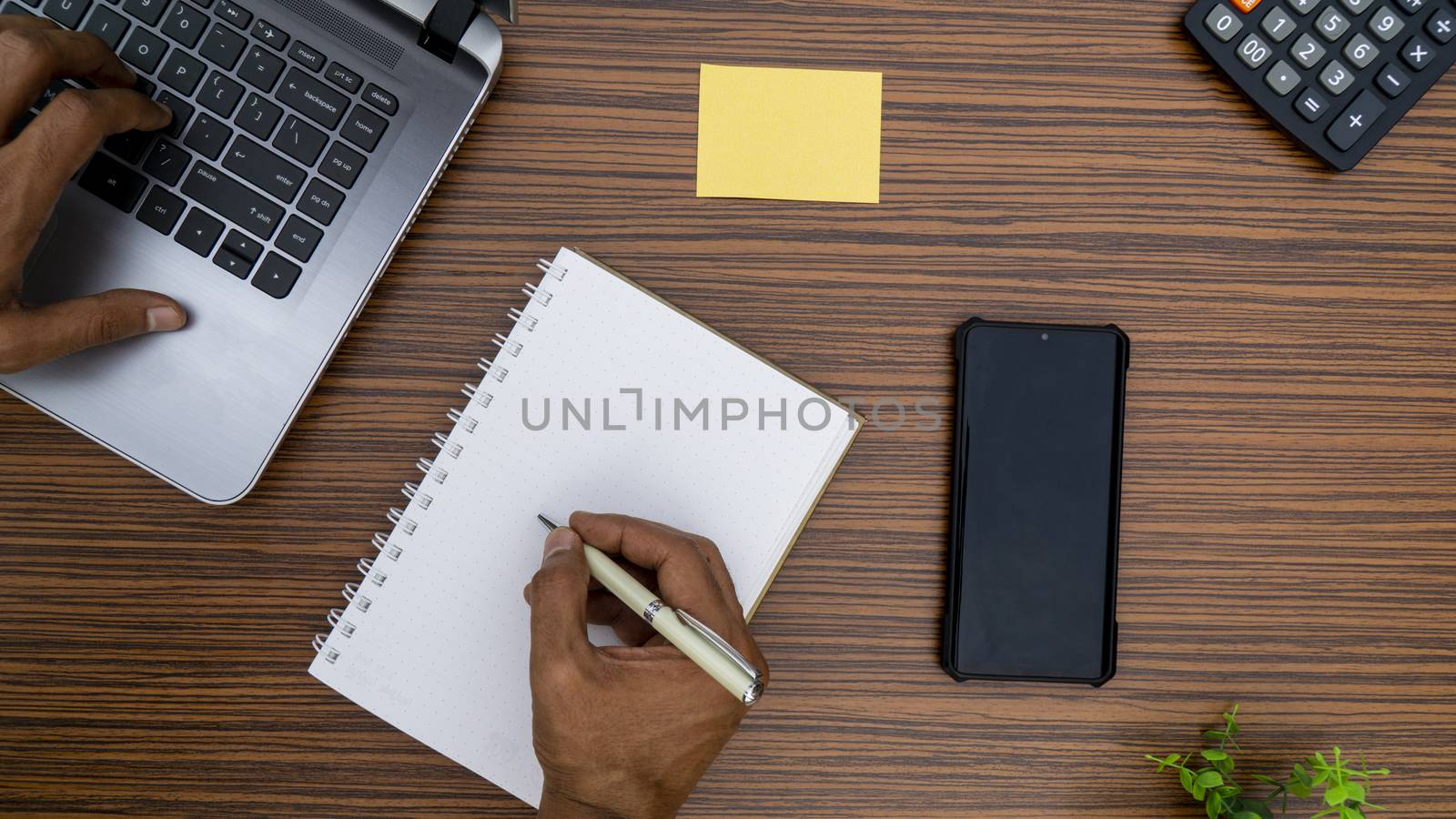 Writing on a notepad and typing on a keyboard while working on a project. A lap top, a mobile, calculator and plant are also on display on this brown striped working table.