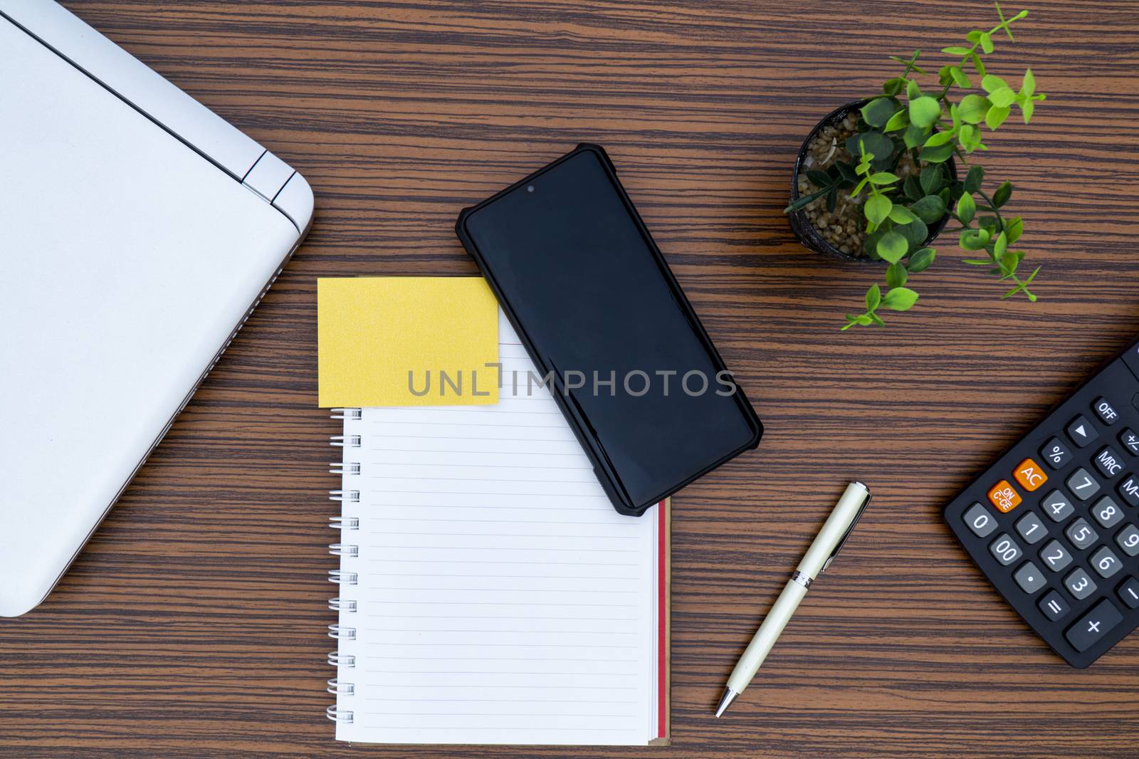 Notepad, sticky note, pen plant, calculator and a lap top on a brown striped zebrawood design table top. Must have objects while working from home during Covid-19