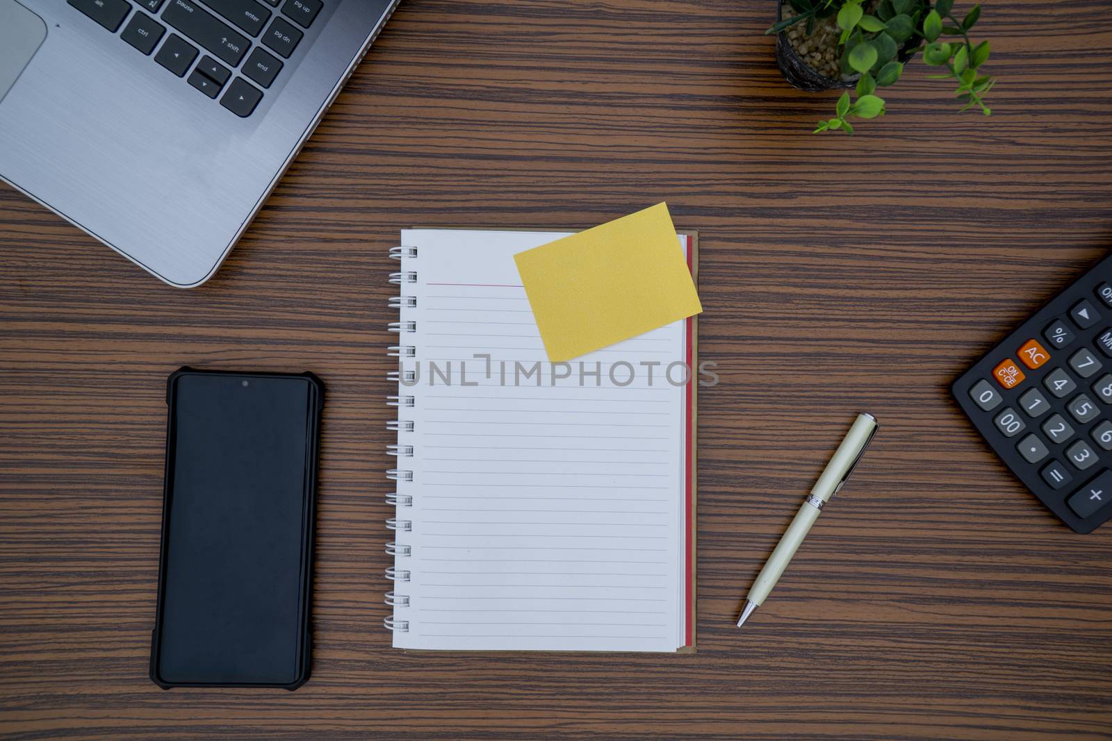 Notepad, sticky note, pen plant, calculator and a lap top on a brown striped zebrawood design table top. Must have objects while working from home during Covid-19 by sonandonures