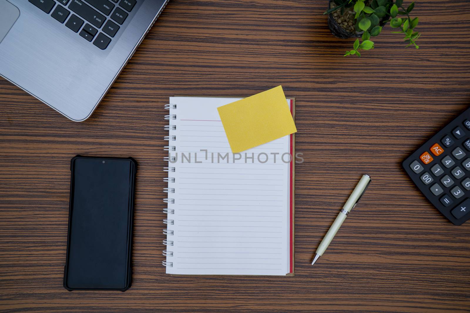 Striped brown working table with note pad, mobile phone, pen, calculator a laptop and table plant on display.