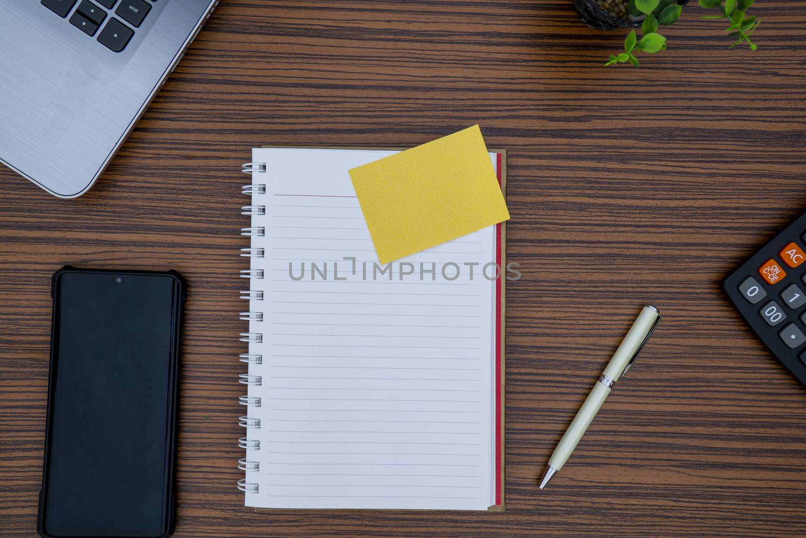 Striped brown working table with note pad, mobile phone, pen, calculator a laptop and table plant on display.