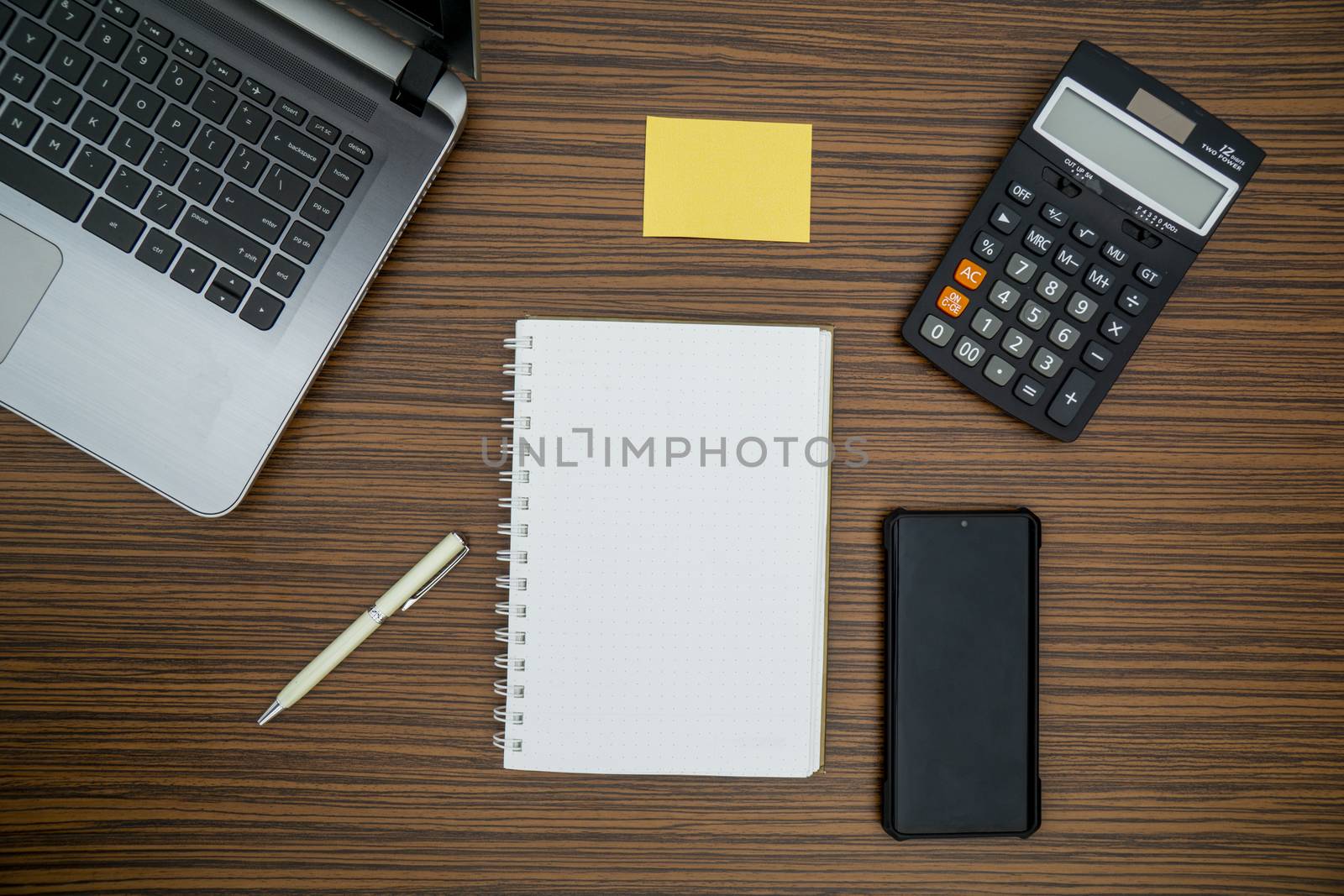 An office table working enviroment. Notepad, sticky note, pen plant, calculator and a lap top on a brown striped zebrawood design table top. Must have objects while working from home during Covid-19