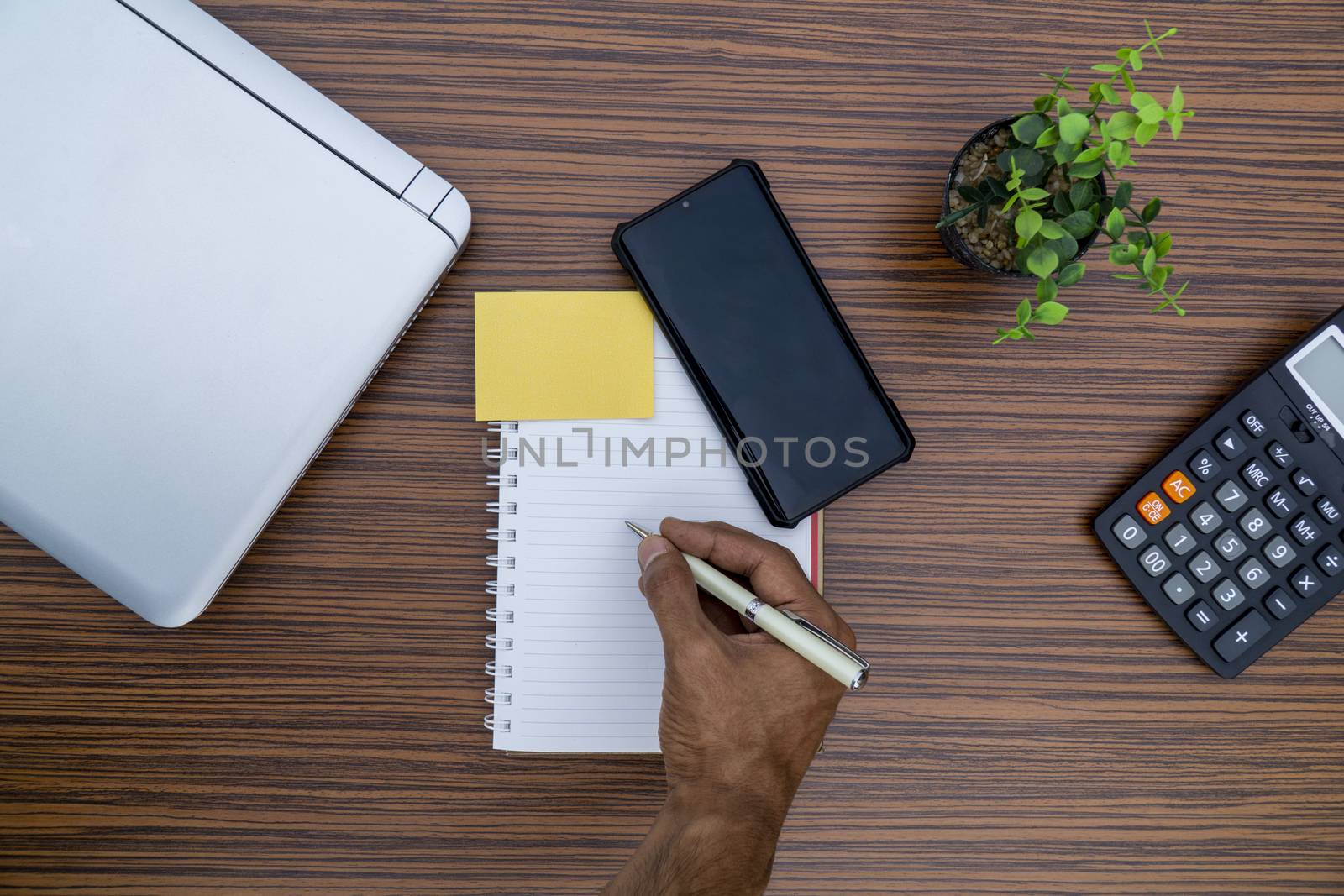 Writing on a notepad while working from home. A lap top, a mobile, calculator and plant are also on display on this brown striped working table.