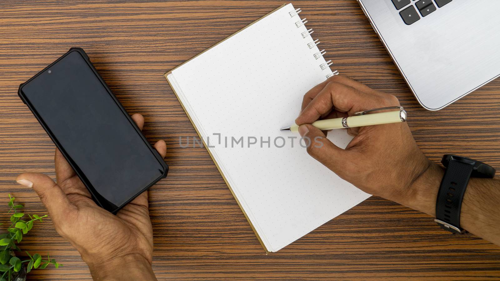 Writing on a notepad while holding a mobile phone working in an office environment. A lap top, a mobile, calculator and plant are also on display on this brown striped working table. by sonandonures