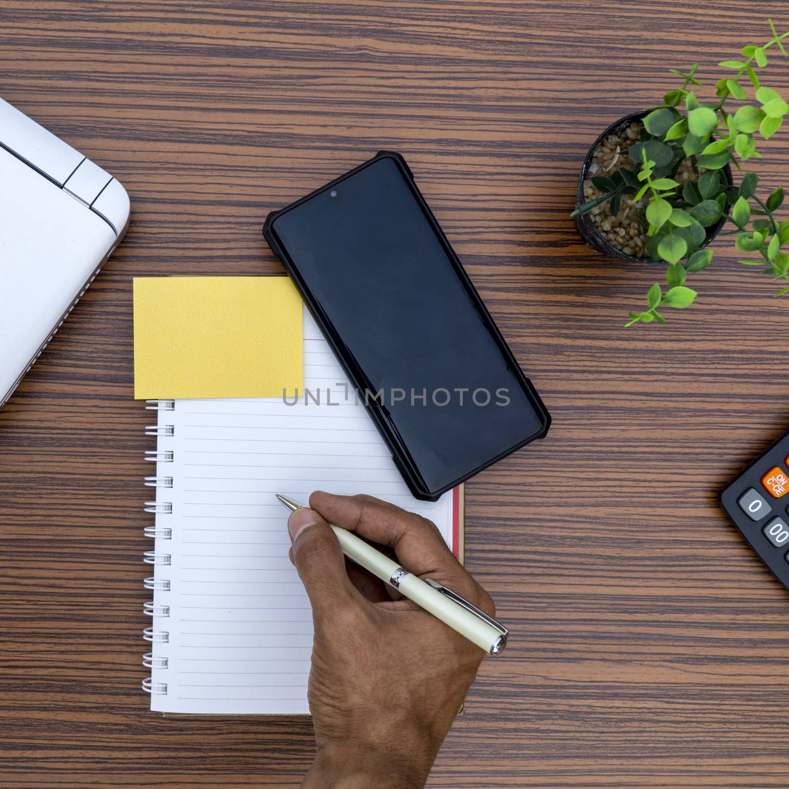 Writing on a notepad while holding a mobile phone working in an office environment. A lap top, a mobile, calculator and plant are also on display on this brown striped working table. by sonandonures