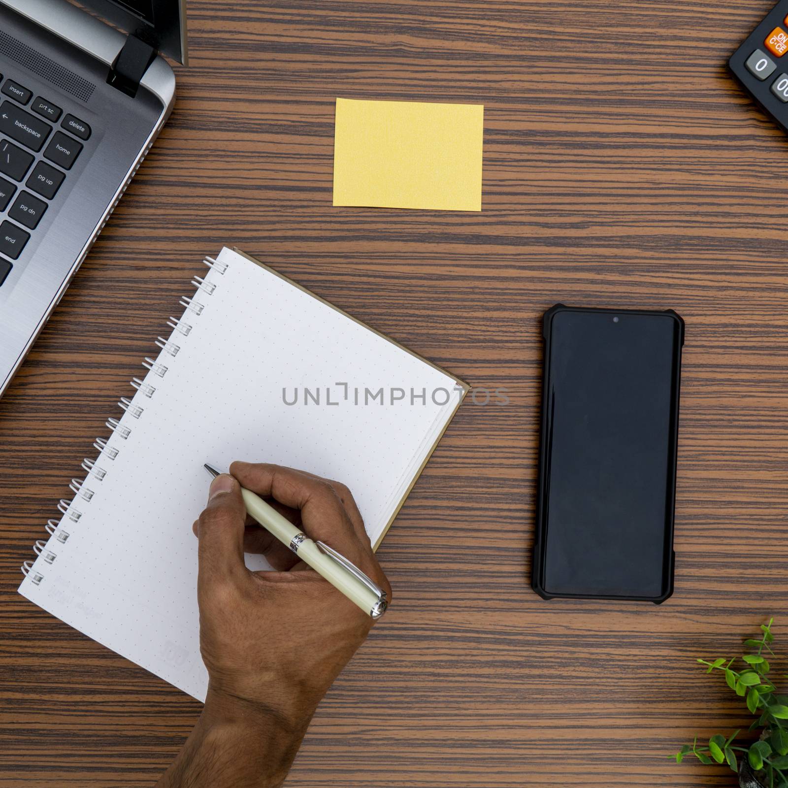 Writing on a notepad while working from home. A lap top, a mobile, calculator and plant are also on display on this brown striped working table.