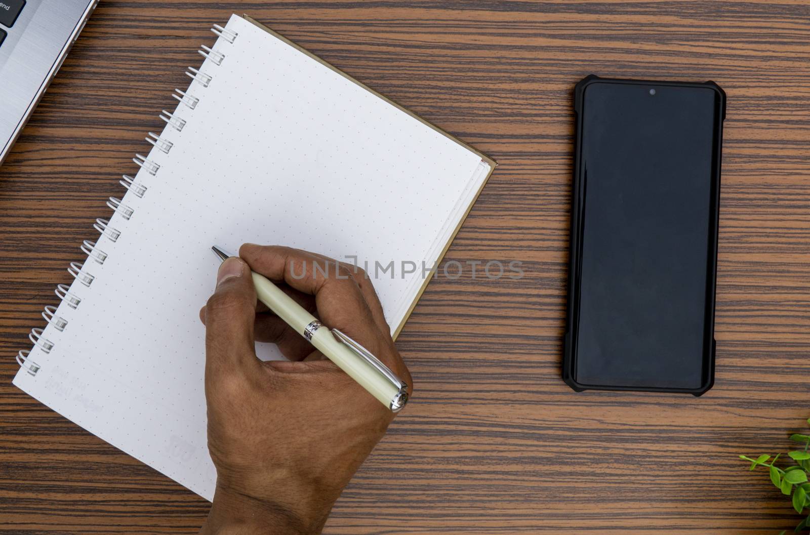 Writing on a notepad while working from home. A lap top, a mobile, calculator and plant are also on display on this brown striped working table. by sonandonures
