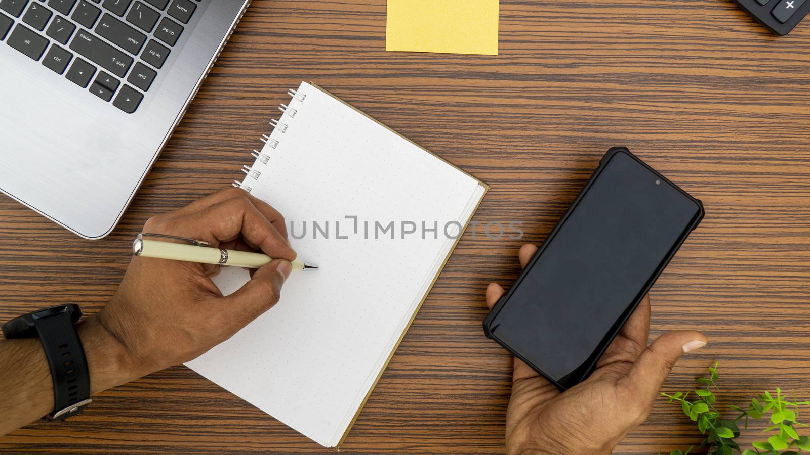 Writing on a notepad while working from home. A lap top, a mobile, calculator and plant are also on display on this brown striped working table. by sonandonures