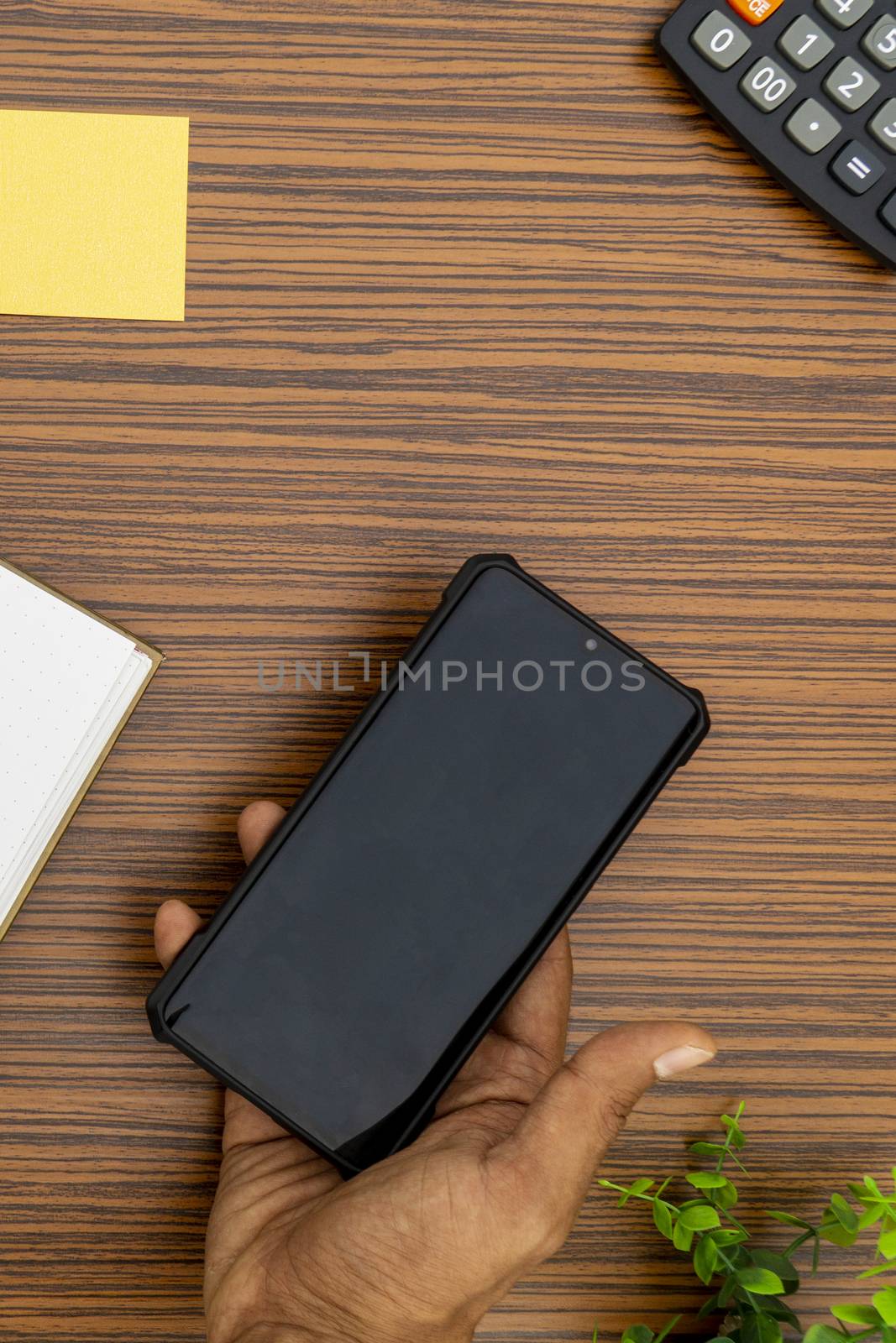 A brown hand holding a mobile phone while working in an office environment. A lap top, a mobile, calculator and plant are also on display on this brown striped working table.