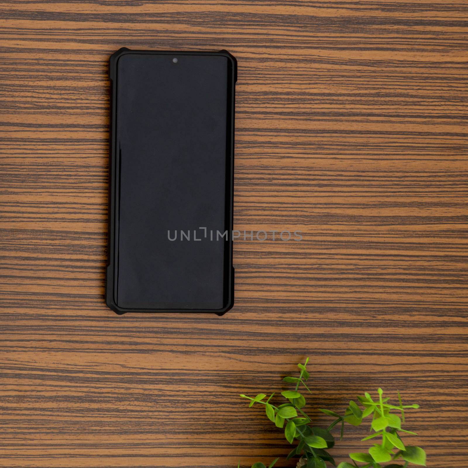 A black mobile phone on a striped brown working table with a calculator and a green plant by sonandonures