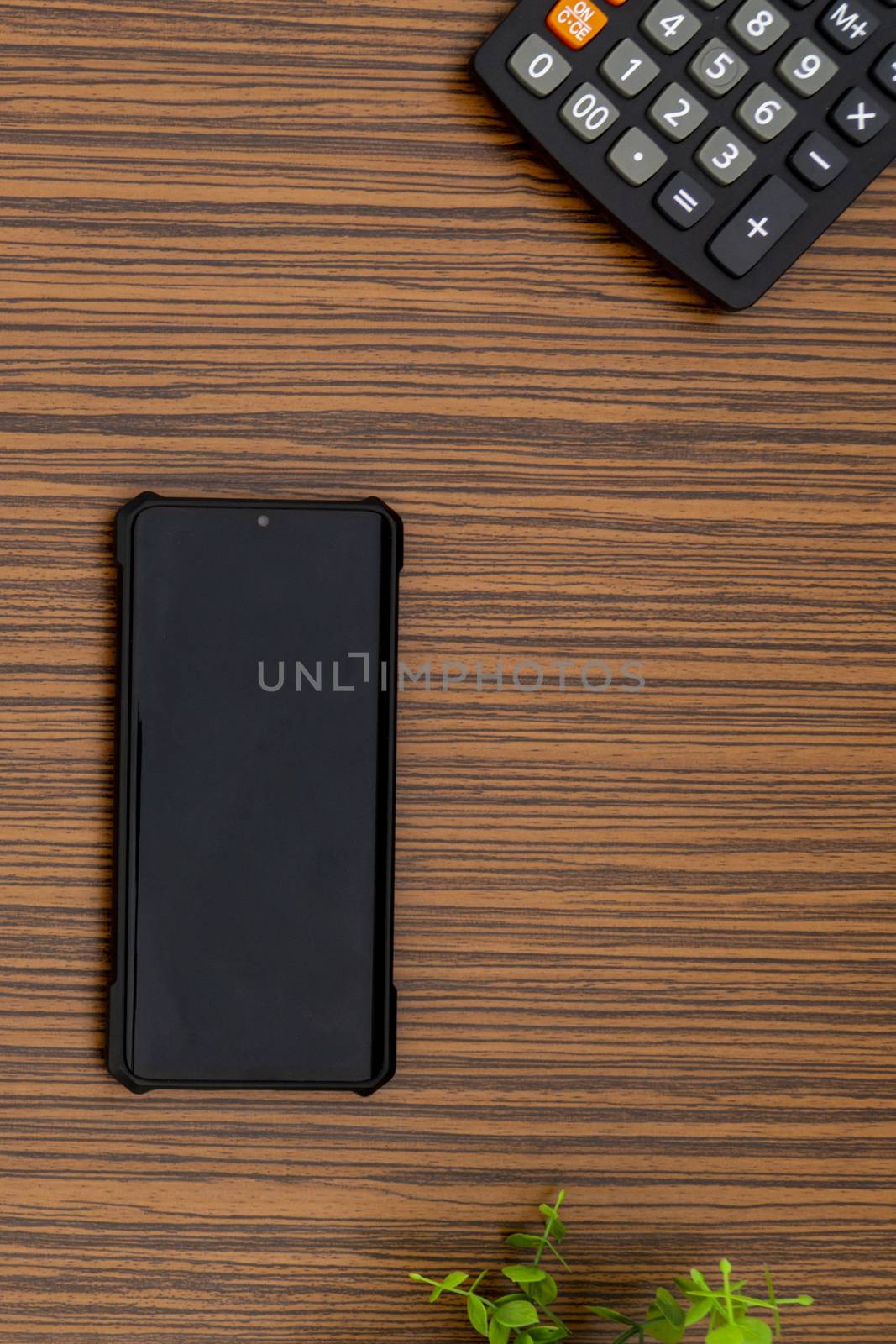 A black mobile phone on a striped brown working table with a calculator and a green plant by sonandonures