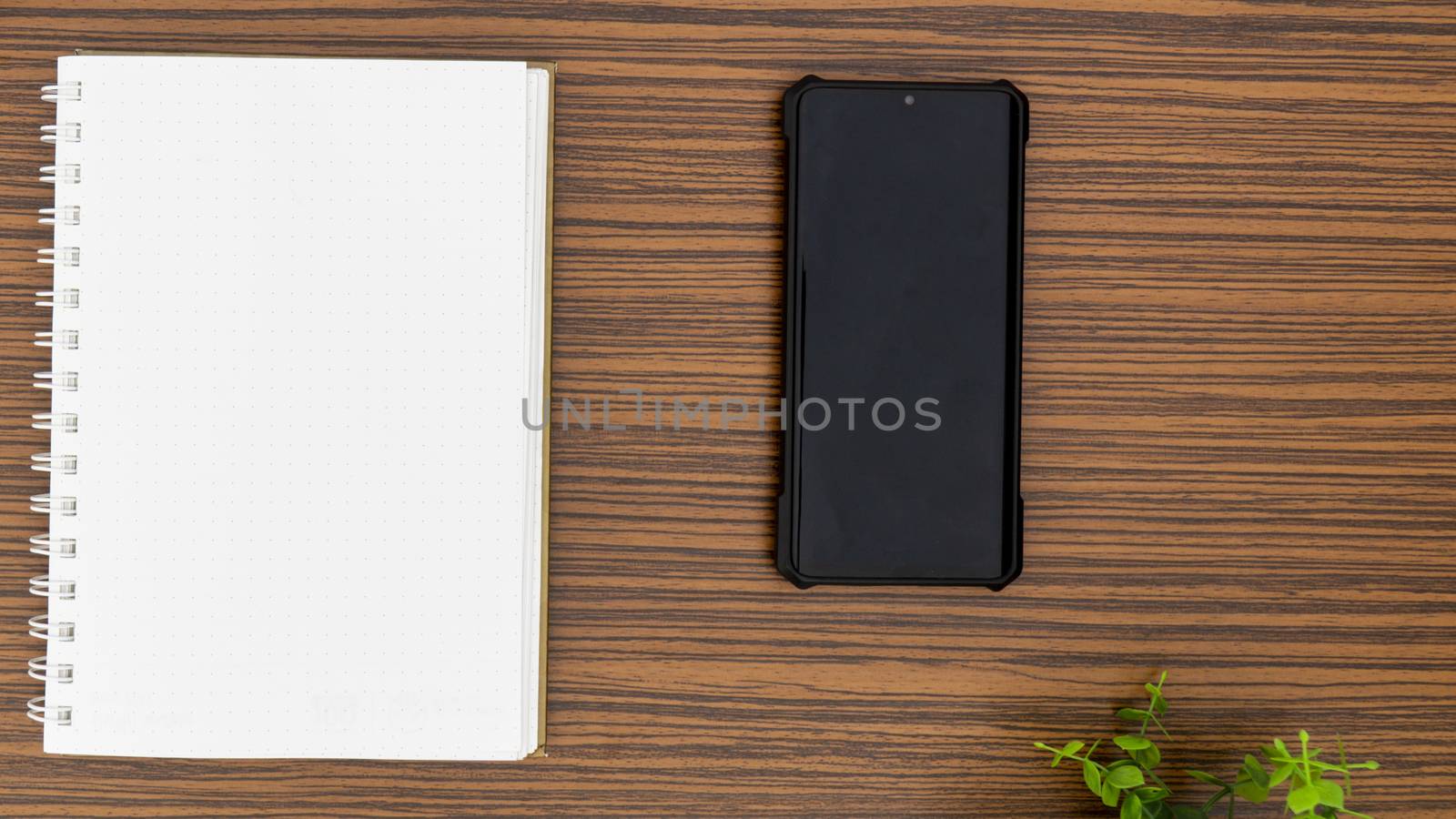 Personal note book and a black mobile phone with a glimpse of green plant on a striped brown office table