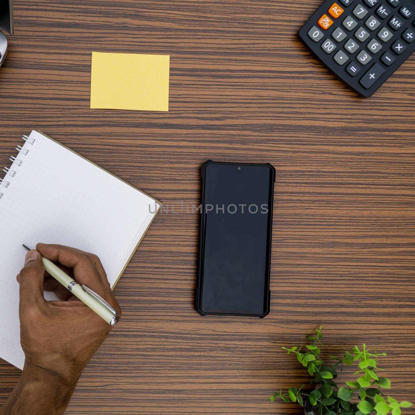 Writing on a notepad while working from home. A lap top, a mobile, calculator and plant are also on display on this brown striped working table. by sonandonures