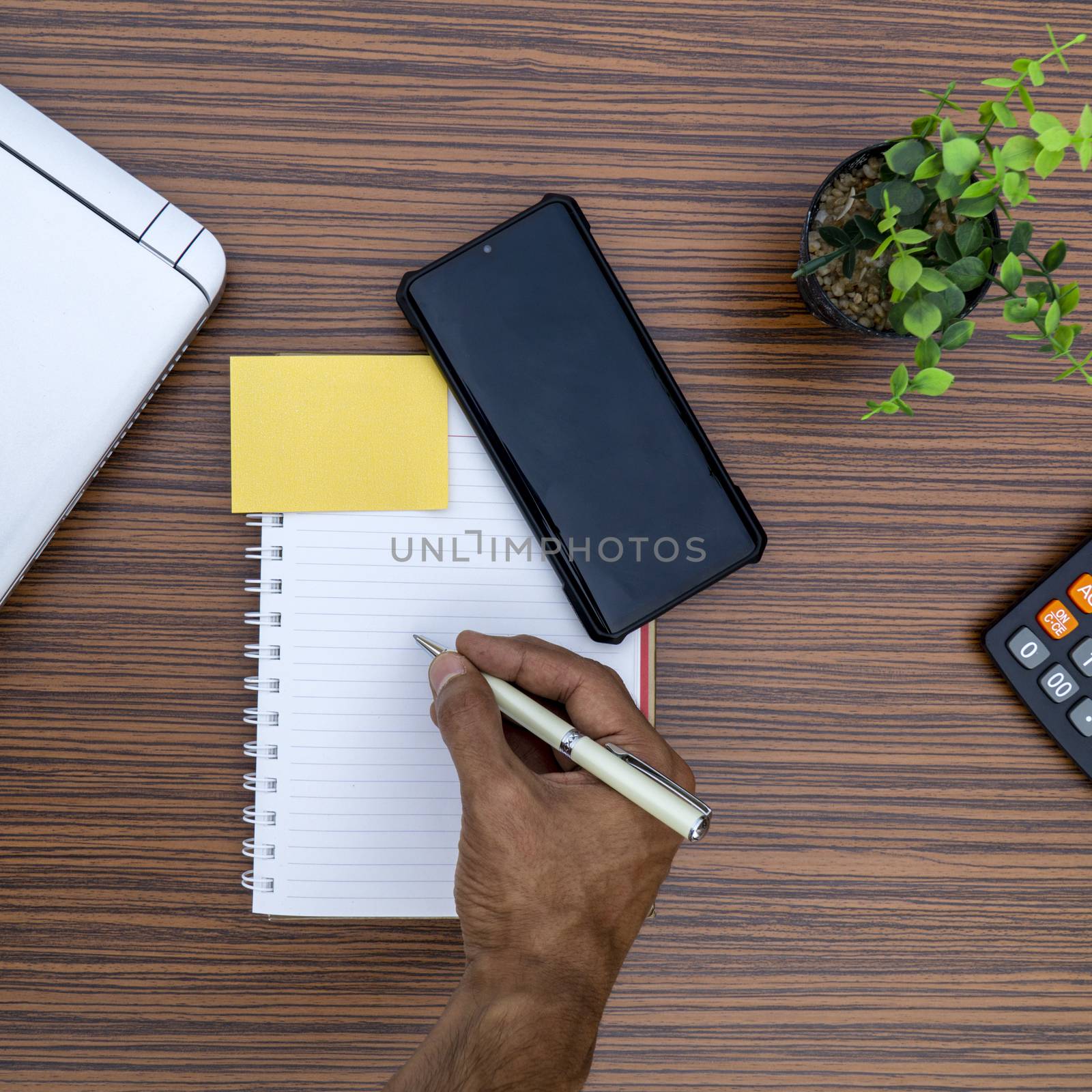 Writing on a notepad while working from home. A lap top, a mobile, calculator and plant are also on display on this brown striped working table. by sonandonures