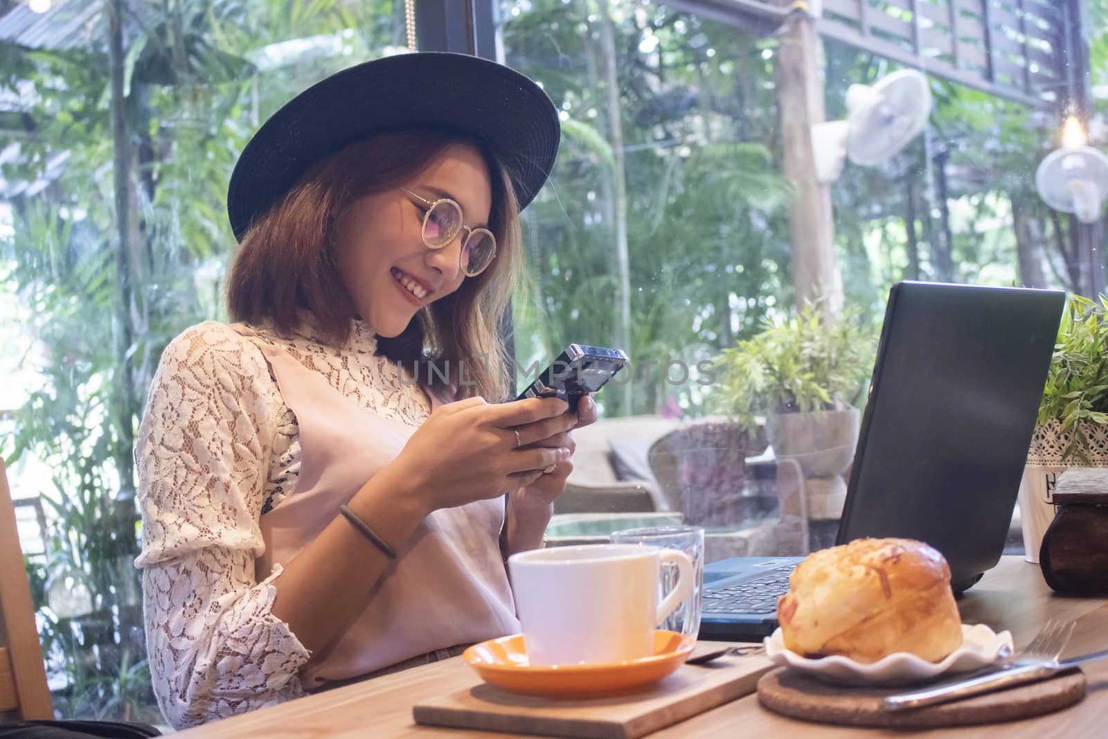 Portrait of a smiling young asian girl holding mobile phone while sitting with laptop computer at a cafe