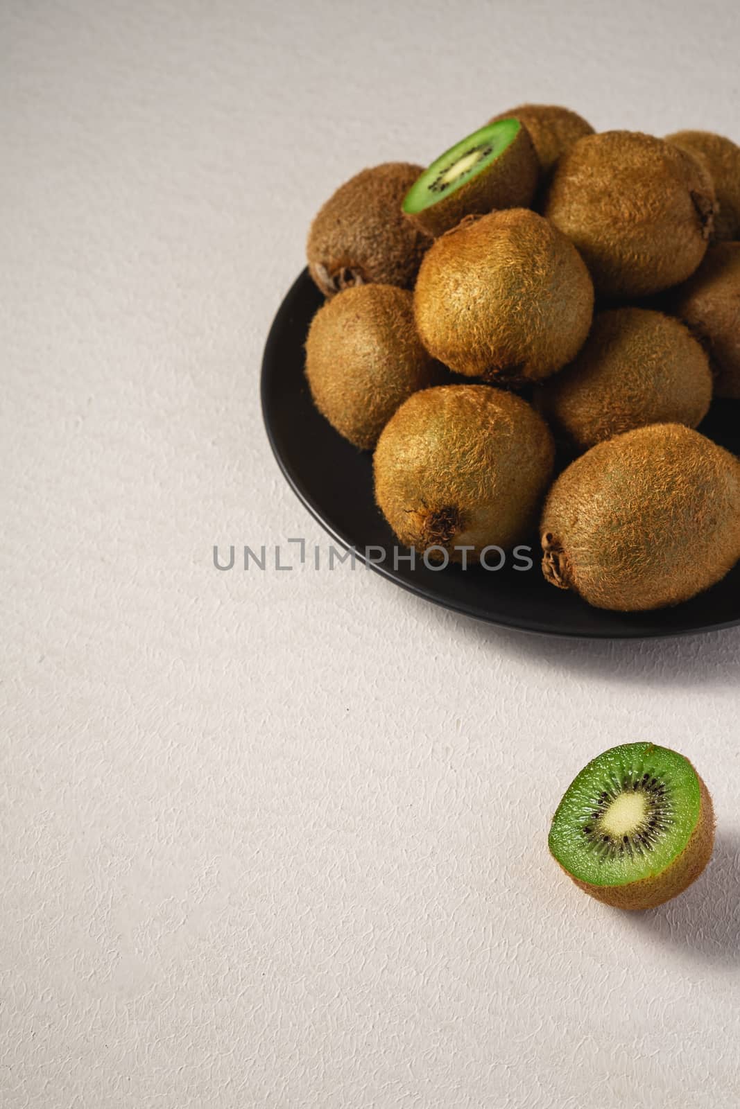 Kiwi fruits half sliced in black plate on vibrant plain white background, copy space, angle view