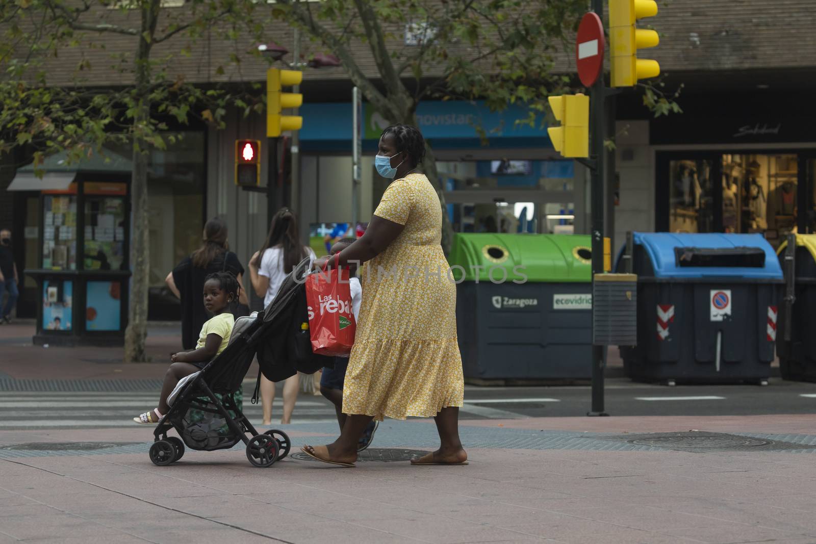 Zaragoza, Spain - August 18, 2020: A black inmigrant woman, with face masks, due to the coronavirus pandemic, pushes a stroller with her child in the central area of Zaragoza.