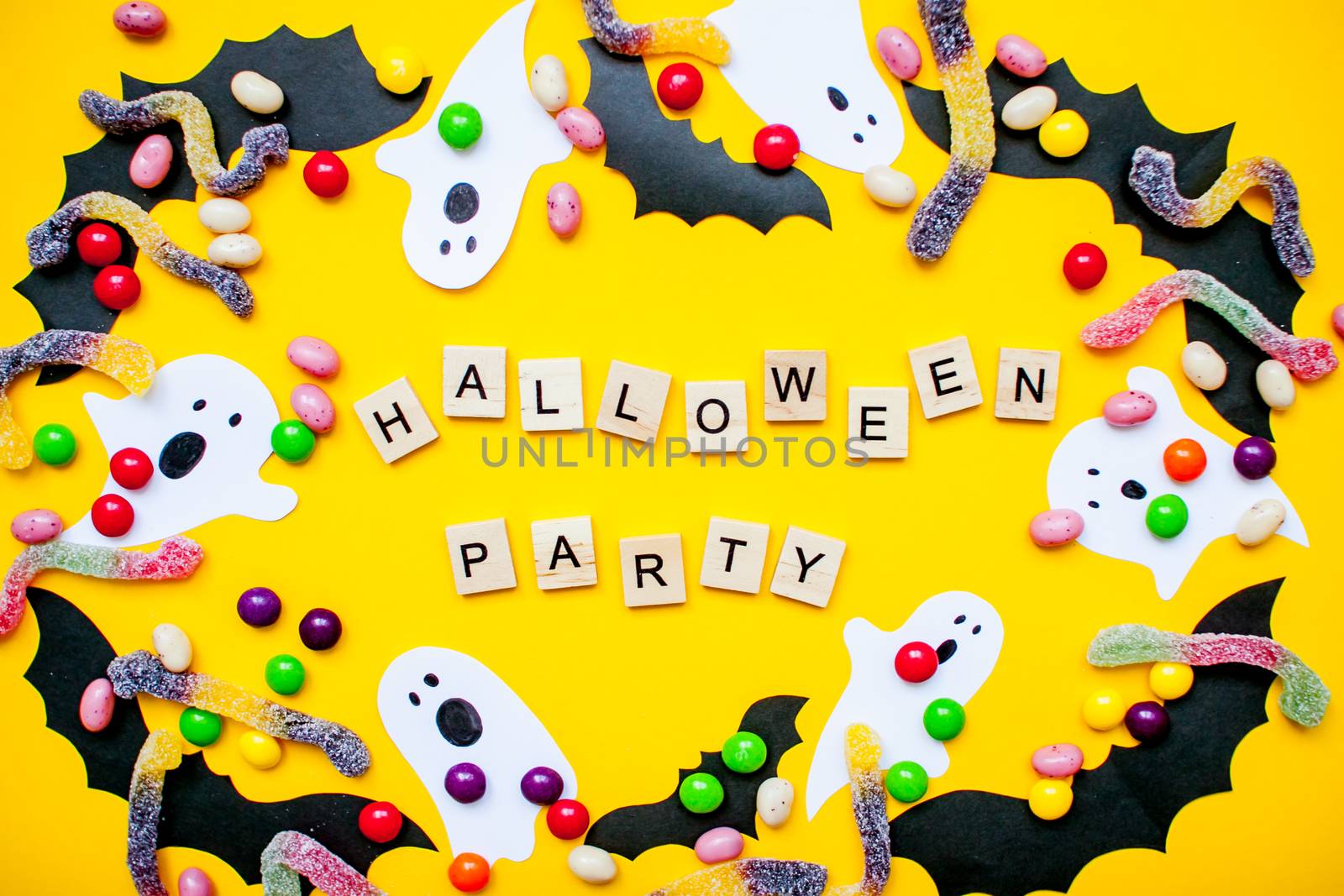 inscription from wooden blocks Halloween party and frame made of paper homemade bats and paper ghosts and multicolored candies and worms from gummy on a bright yellow background
