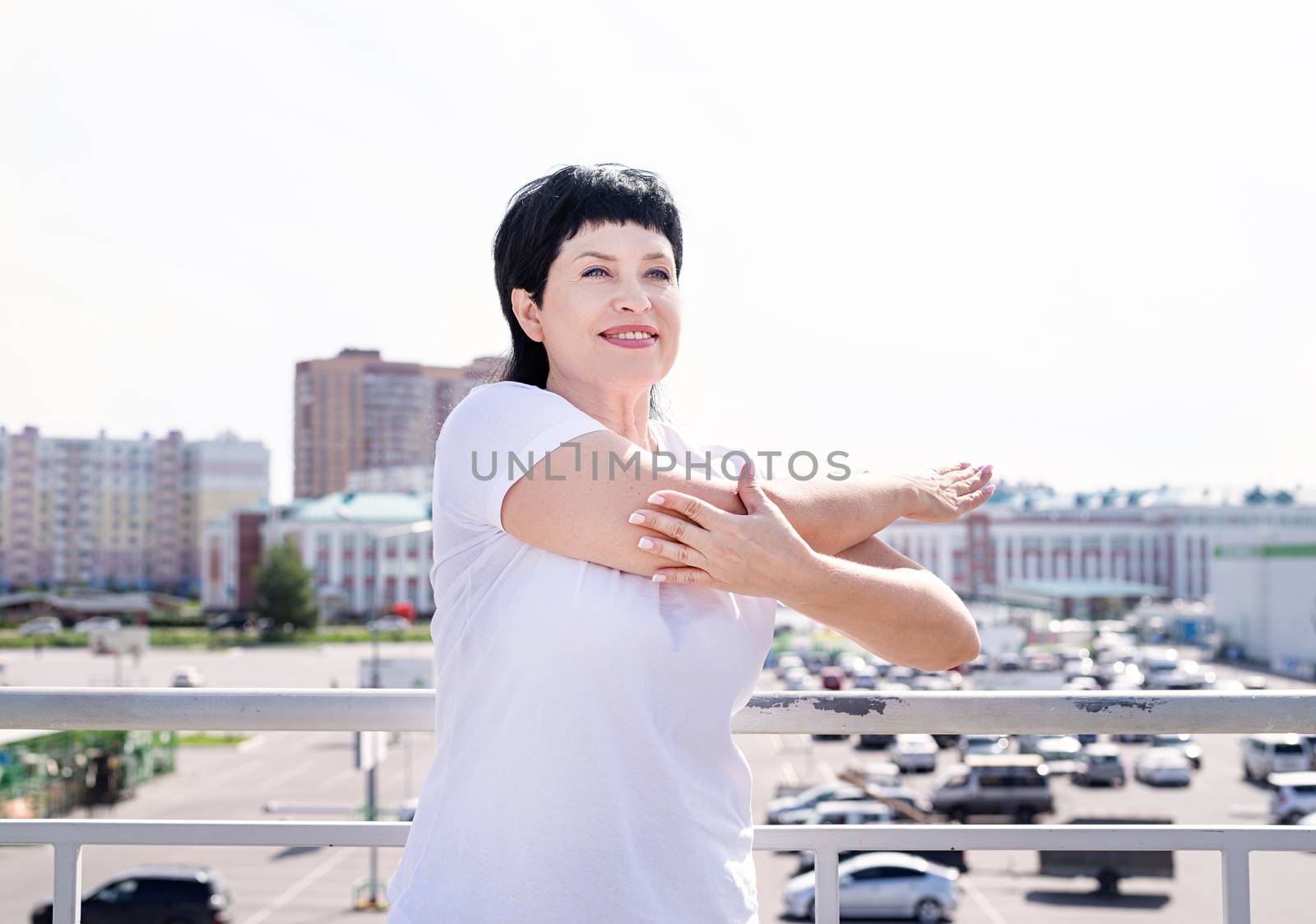 Smiling senior woman doing stretching outdoors on urban background by Desperada