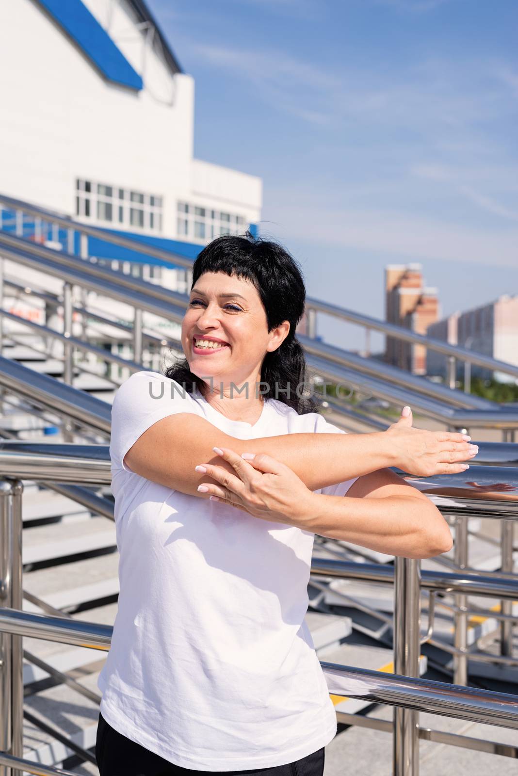 Smiling senior woman doing stretching outdoors on urban background by Desperada